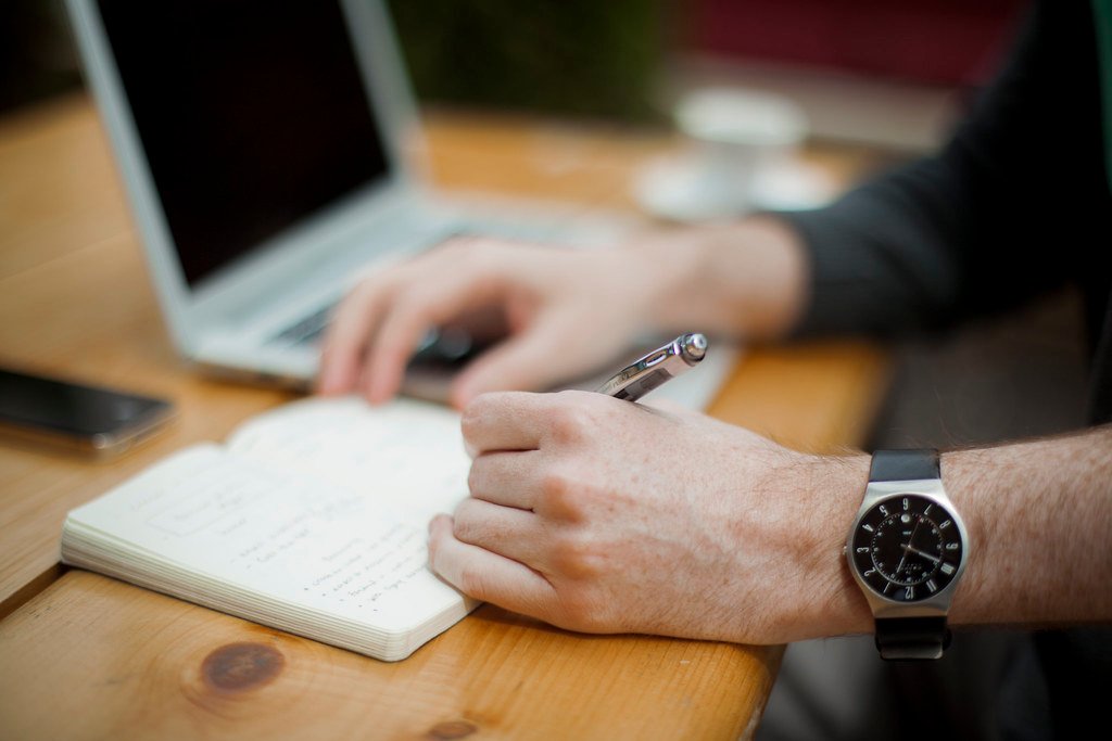 A notebook and pen open on a desk next to a laptop, used for drafting page descriptions